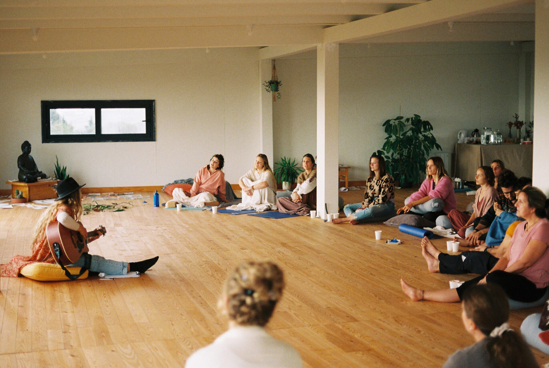 A singer is singing and playing the guitar; the audience is watching her. The concert is in the cozy yoga shala.